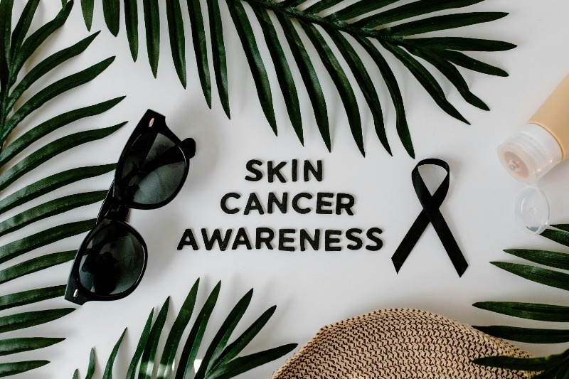 Picture that say Skin Cancer Awareness with Cancer ribbon, sunglasses, sun screen and the brim of a hat with palm leaves.